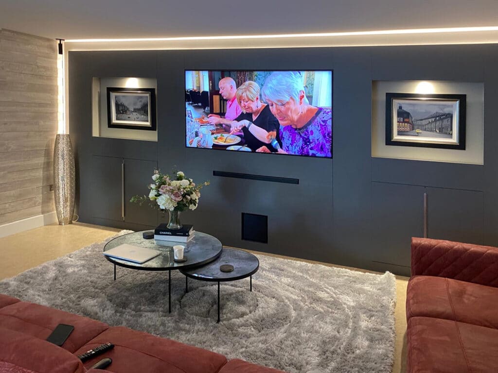 Bespoke media wall by Hawker's with a wall-mounted TV, soundbar, recessed framed-art niches and LED accent lighting on a dark panelled wall